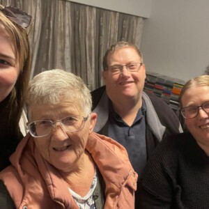 John's family - including his elderly mother - gather for a joyful photo, smiling and standing close together. There are three women in the front and two men in the back.